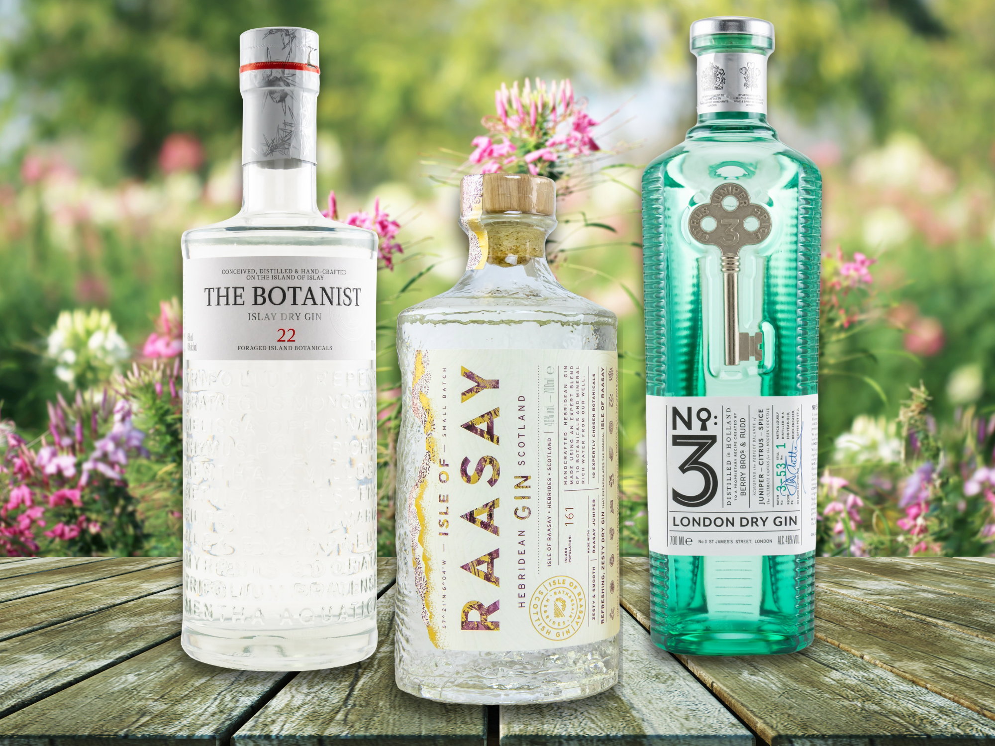 The Botanist Islay Dry Gin 22 in klarer Flasche, Raasay Hebridean Gin in edler Flasche und No. 3 London Dry Gin in blaugrüner Flasche, umgeben von blühenden Pflanzen.