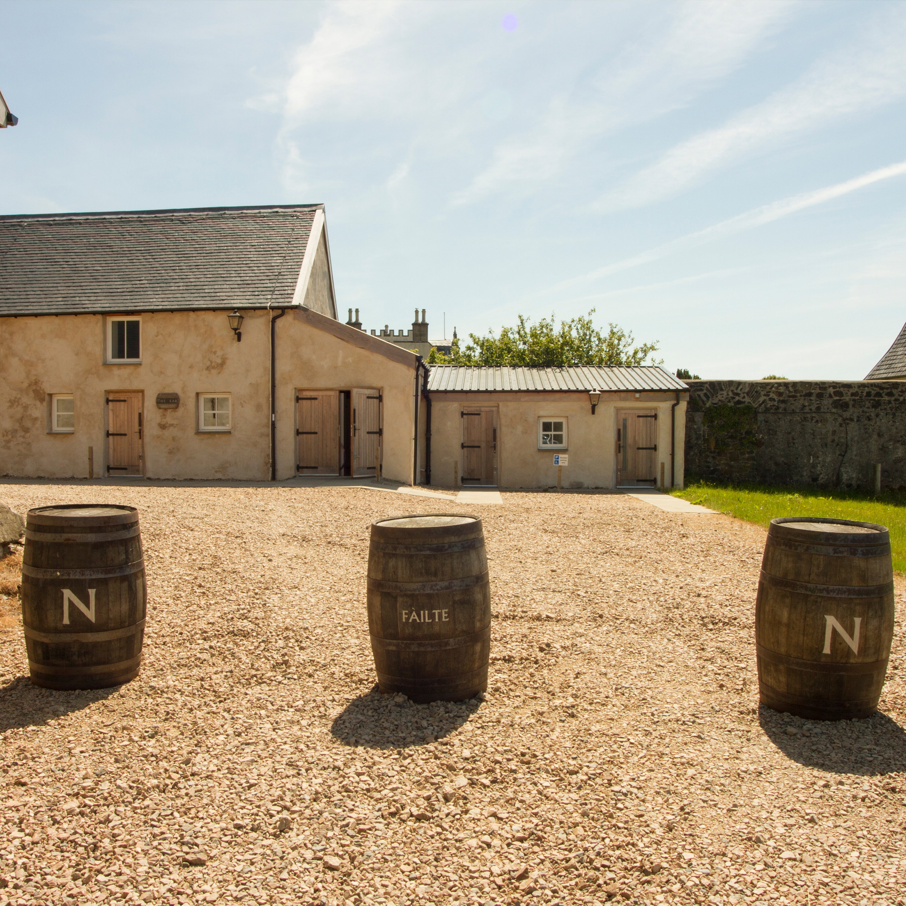 Historische Destillerie-Hofanlage mit drei großen Whiskyfässern als Kompass-Nordmarkierung auf Kiesboden vor rustikalen Steingebäuden