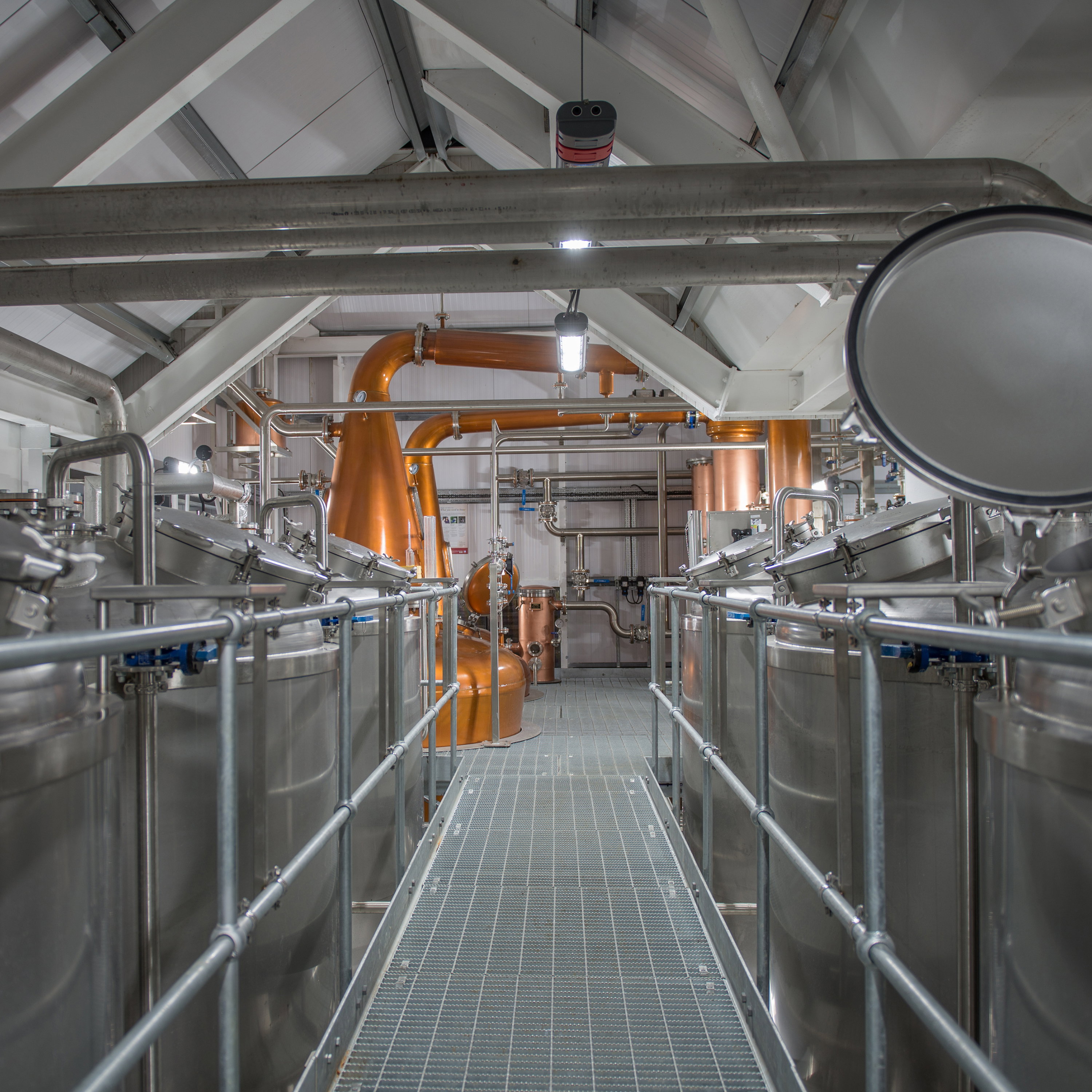 Moderne Kupferbrennblasen und Edelstahltanks in einer Premium-Whisky-Destillerie mit industriellem Rohrleitungssystem und Gitterboden für handwerkliche Spirituosenherstellung