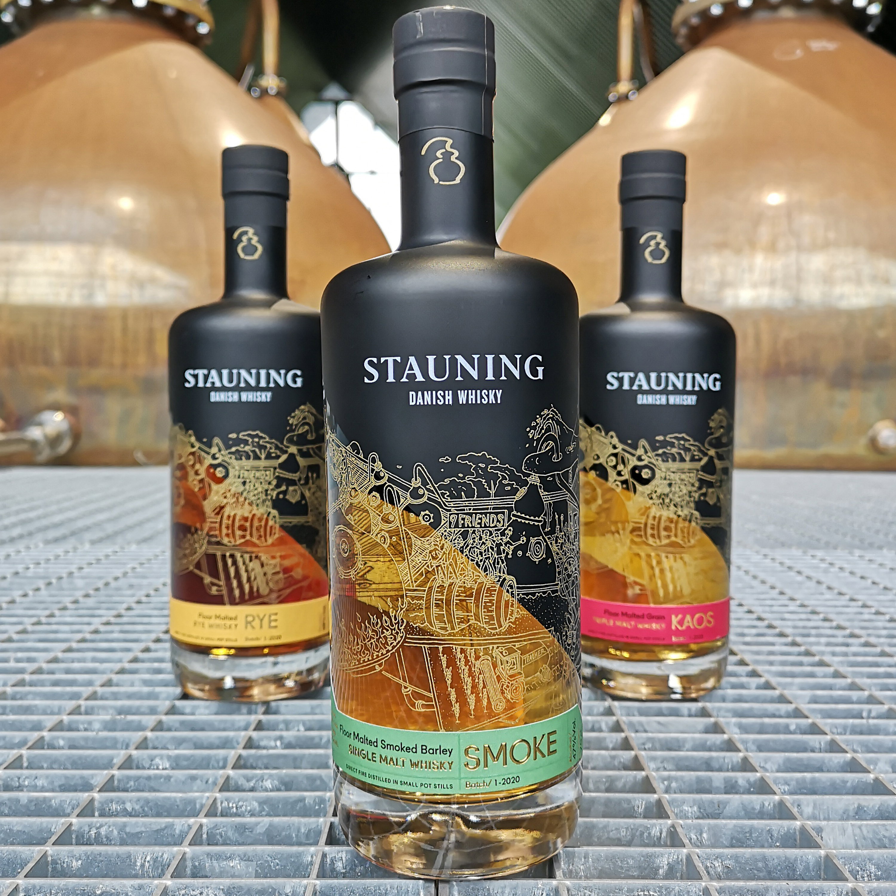 Stauning Danish Whisky Rye, Smoke und Kaos Editionen als Trio mit aufwendigen gold-schwarzen Etiketten vor Kupferbrennblasen-Destilleriehintergrund