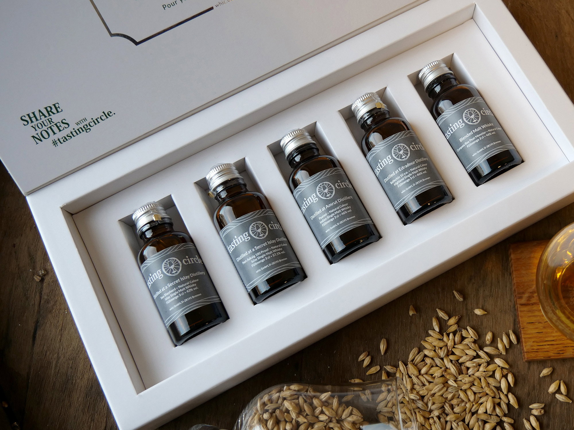 Tasting Circle Whisky-Discovery-Set mit 5 Single Malt Whiskys aus Secret Islay, Amrut, Edradour, Blended Malt in weißer Präsentationsbox auf rustikaler Holzoberfläche mit Getreidekörnern
