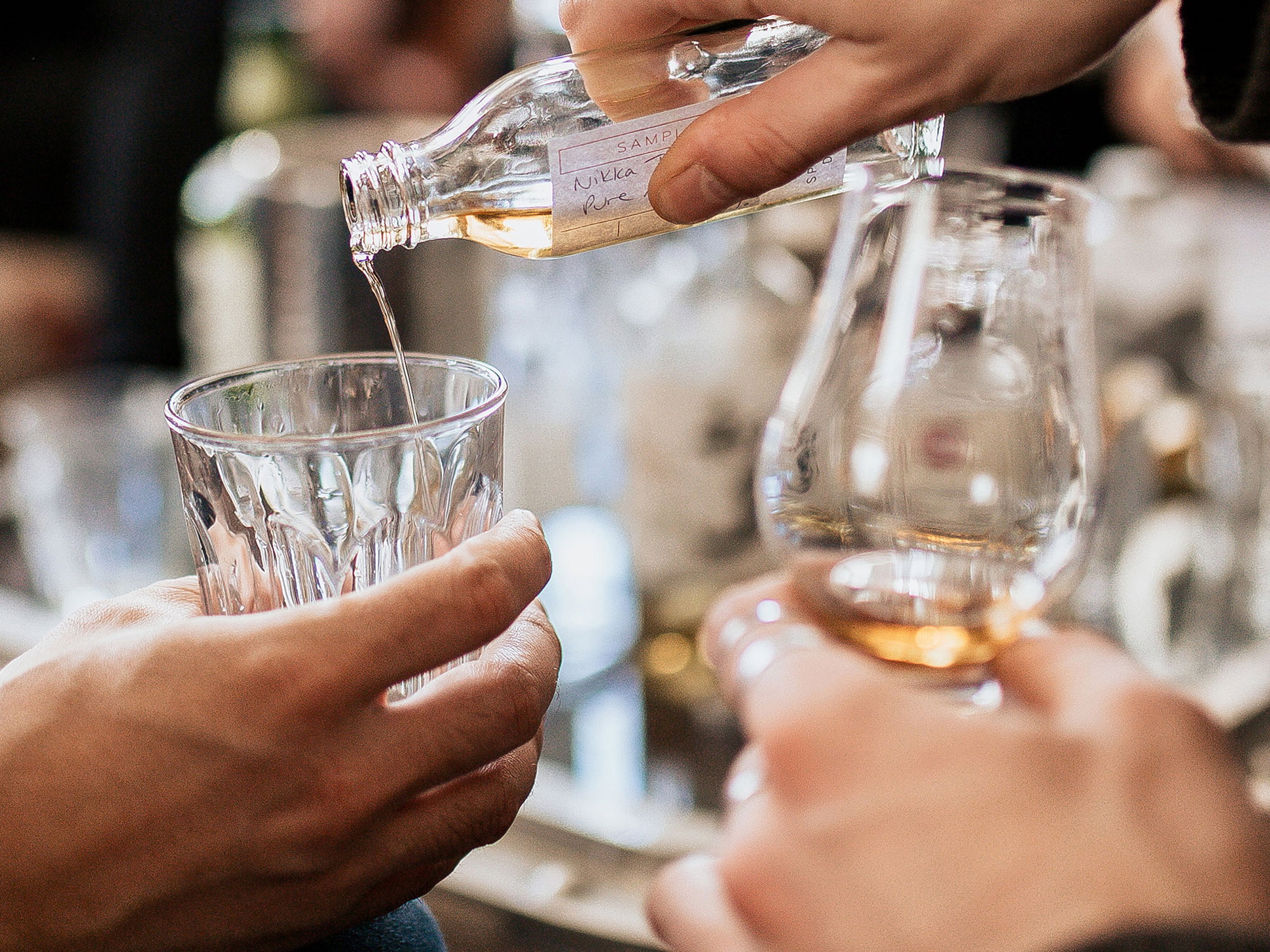 Premium-Whisky-Verkostungsszene mit Einschenken einer klaren Spirituose aus Miniaturprobe-Flasche in kristallklares Nosing-Glas, im Hintergrund Weinglas mit goldfarbenem Whisky